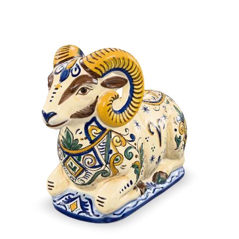Ariete in maiolica accucciato, con grandi corna arricciate in zafferano e il vello bianco dipinto a mano con decori geometrici blu cobalto.