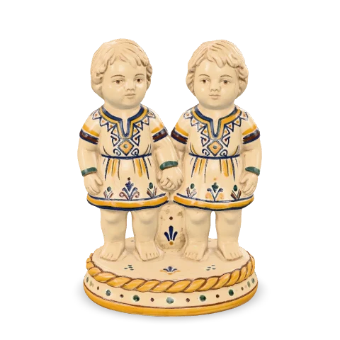 Due bambini in maiolica in piedi uno accanto all’altro, per mano, con tuniche bianche decorate in cobalto e zafferano, su una stessa base tonda.