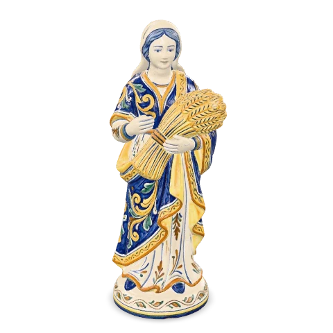 Fanciulla in maiolica in piedi, in una lunga veste decorata di blu cobalto, che stringe fra le braccia un covone di grano dorato.